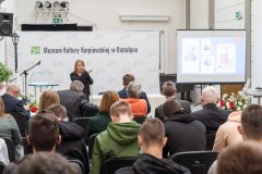 Uczestnicy konferencji słuchają przemówienia prelegentki stojącej przed widownią. W tle ścianka z napisem Muzeum Kultury Kurpiowskiej w Ostrołęce oraz logotypami Muzeum. Z prawej strony ekran z wyświetloną prezentacją.