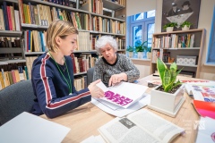 Dwie kobiety przy stole oglądają wycinanki kurpiowskie. W tle regały biblioteczne.