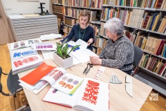 Dwie kobiety siedzące przy stole w bibliotece oglądające wycinanki kurpiowskie.