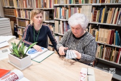 Dwie kobiety siedzą przy stole, jedna z nich wykonuje wycinankę. W tle regał biblioteczny.