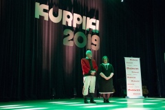 Na scenie stoi mężczyzna w stroju kurpiowskim oraz kobieta, obok stoi rollup z logami marki Mazowsze. W tle duży napis KURPIKI 2019.