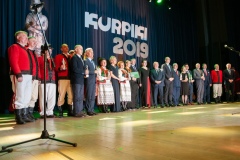 Na scenie stoi w rzędzie ponad dwadzieścia osób, niektórzy w strojach kurpiowskich. W tle duży napis KURPIKI 2019