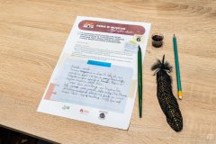 Na stole leży karta edukacyjna z pismem drukowanym i ręcznym. Obok leży pióro wieczne, pióro ptasie oraz ołówek.