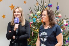 Dwie kobiety, jedna z nich mówi do mikrofonu. W tle choinka świąteczna, dalej ściana z ozdobami w postaci gwiazdek