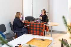 Dwie kobiety siedzą po przeciwnych stronach stołu, zwrócone ku sobie, każda trzyma mikrofon. Jedna z kobiet ma założone słuchawki