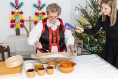 Kobieta w kurpiowskim stroju przygotowuje w kopońce ciasto. Przed nia na stole stoją naczynia gliniane, obok stolnica z sitem do mąki. W większym naczyniu  starta marchew. Z prawej strony stoi kobieta z mikrofonem.