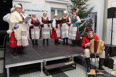 Na scenie stoi kilka kobiet w strojach kurpiowskich. Pod sceną siedzi mężczyzna grający na harmonii pedałowej.