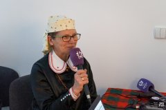 Przy stole siedzi kobieta w kurpiowskim stroju i trzyma mikrofon