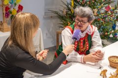 Dwie kobiety siedzą przy stole. Jedna z kobiet trzyma mikrofon rozmawiając z drugą, która jest w kuprpiowskim stroju. W tle choinka bożonarodzeniowa.