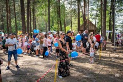 Kilkadziesiąt osób na terenie Zagrody Kurpiowskiej, wokół drzewa, w tle kurpiowska chata