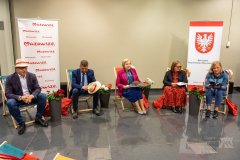 Na holu w mauzoleum Muzeum Kultury Kurpiowskie w Ostrołęce siedzą trzy kobiety i dwóch mężczyzn na leżakach. Jeden mężczyzna ma kapelusz na głowie a drugi trzyma w ręku. Kobieta po prawej stronie czyta książkę. W tlerbem województwa mazowieckiego przedstawiający w polu czerwonym białego orła, o dziobie i szponach żółtych, po prawej stronie stoi baner z he. A po lewej stronie stoi baner biały z czerwonymi napisami Mazowsze serce Polski.