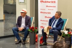 Dwóch mężczyzn siedzących na leżakach, mężczyzna z lewej strony ma okulary i kapelusz na głowie, drugi trzyma go w ręce. Obok każdego stoją kwiaty w doniczkach a za nimi stoi biały baner z czerwonymi napisami Mazowsze serce Polski. 