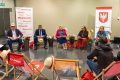 Grupa osób siedzi na leżakach  w półkolu na holu mauzoleum w Muzeum Kultury Kurpiowskiej. Mężczyzna po lewej stronie ma kapelusz na głowie a drugi trzyma w ręku, dalej siedzą trzy kobiety i mężczyzna odwrócony plecami. W tle po prawej stronie stoi  baner Marszałek Województwa Mazowieckiego. A po lewej stronie stoi baner z napisem Mazowsze serce Poski.  
