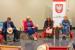 Trzy kobiety i jeden mężczyzna siedzą na leżakach, między leżakami stoją kwiaty biało czerwone. W tle baner z herbem województwa mazowieckiego przedstawiający w czerwonym polu białego orła, o dziobie i szponach żółtych. 