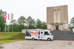 Mazowiecki kamper z napisami Mazo podróże stoi przed pomnikiem Mauzoleum na Fortach w Ostrołęce. po lewej stronie na tle zielonych drzew powiewają trzy maszty z flagami   