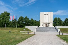 Pomnik Mauzoleum, wokół zielony teren, z lewej strony Pomnika trzy flagi na maszcie (od lewej flaga Mazowsza, Polski, Unii Europejskiej). W tle drzewa. Widok w słoneczny dzień.
