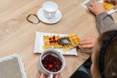 Przy stole, dłonie trzymające filiżankę z czerwonymi konfiturami, nakładające konfitury na gofry, obok mniejsza, pusta filiżanka na spodku, z lewej strony fragment obszytej koronkową taśmą serwetki