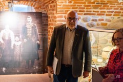 W zabytkowych piwnicach przemawia mężczyzna, obok stoi kobieta. W tle na ekspozycji wielkie stare zdjęcie przedstawiające kobietę i mężczyznę oraz dziecko