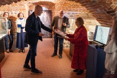 Goście na wernisażu w zabytkowych piwnicach. Mężczyzna podaje kobiecie książkę.