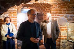 Goście na wernisażu w zabytkowych piwnicach. Mężczyzna przemawia, w tle stoi kobieta i inny mężczyzna