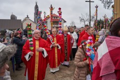 Księża w czerwonych ornatach idący do kościoła z palmami wielkanocnymi. Wśród nich Biskup. Wokół tłum z palmami oczekujący na rozpoczęcie procesji.