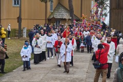 Procesja z palmami wielkanocnymi, w tle drewniany kościół. Pierwsi idą księża i ministranci w komżach. Mężczyzna z przodu trzyma pionowo krzyż.