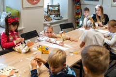 Dzieci siedząc przy stole wykonują z kartonu lampiony w kształcie budynku Muzeum. Z prawej strony siedząca przy stole opiekunka grupy