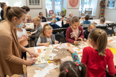 Dzieci siedząc przy stole wykonują z kartonu lampiony w kształcie budynku Muzeum. Z lewej strony pracownica Muzeum pomaga dzieciom w wykonaniu lampionów