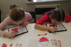 Dwie dziewczynki wykonują techniką malowania na szkle biało-czerwone obrazki inspirowane wycinanką kurpiowską