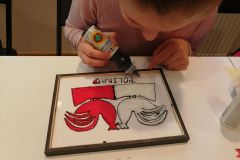 Dziewczynka wykonuje techniką malowania na szkle, biało-czerwony obrazek inspirowany wycinanką kurpiowską w formie dwóch kogutów