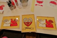 Na stole leżą trzy obrazki oprawione w drewniane ramki, wykonane techniką malowania na szkle, przedstawiające biało-czerwone wycinanki kurpiowskie (dwa zewnętrzne obrazki przedstawiają koguty, obrazek środkowy przedstawia serce). Za obrazkami , na stole leżą przybory i wzorniki