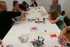 Dzieci i dorośli siedząc przy wspólnym, długim stole, wykonują techniką malowania na szkle biało-czerwone obrazki inspirowane wycinanką kurpiowską