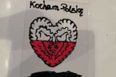 Biało-czerwone serce inspirowane wycinanką kurpiowską, wykonane techniką malowania na szkle. Nad sercem napis „Kocham Polskę”