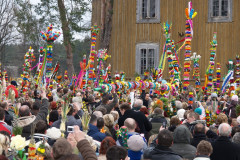  tłum niosący w procesji wysokie kurpiowskie palmy wielkanocne w tle fragment starego drewnianego kościoła jedna z dłuższych palm niesiona jest poziomo