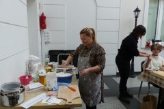 Kobieta w kuchennym fartuchu stoi przy stole, na którym leżą przybory kuchenne i produkty żywnościowe, miesza mąkę w misce. Dalej, z prawej strony, przy stole stoi kobieta i siedzi dziecko.