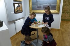Kilkoro dzieci stojąc przy okrągłym stoliku lub siedząc na podłodze rozwiązują karty edukacyjne. W tle, na ścianie mniejsze i duże obrazy w ramach