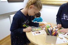 Dziewczynka stoi przy okrągłym stoliku i kredką koloruje obrazek w książeczce. Widać, że w pobliżu są też inne dzieci. W tle na ścianie  widać fragmenty wiszących obrazów.