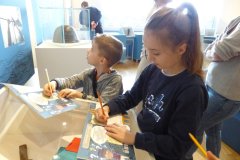 Dziewczynka i chłopiec rysują/piszą w kolorowych książeczkach, położonych na gablocie. W tle inne osoby, gabloty, plansze wystawy.