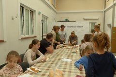 Na muzealnym dziedzińcu, wokół długiego stołu siedzą lub stoją dzieci i dorośli. Na stole leżą tekturowe tacki z fafernuchami. W tle ścianka z nazwą i logami Muzeum Kultury Kurpiowskiej w Ostrołęce