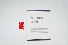 Na ścianie Opis wystawy „W lustrze uliczek”. Obok naklejony czerwony duszek.