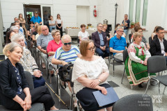 Uczestnicy wernisażu siedzący na widowni na muzealnym dziedzińcu. Niektóre osoby stoją. W tle wejście na dziedziniec.