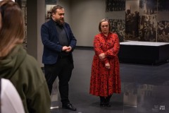 Mężczyzna i kobieta stoją w sali wystawowej. W tle na ścianie fragment ekspozycji w postaci dużych, starych fotografii