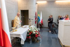Ksiądz odprawiający mszę w krypcie Pomnika Mauzoleum oraz uczestnicy mszy. Z prawej strony fragment sarkofagu. Z lewej fragment flagi Polski.  W  tle w stojaku flagi: Polski, Województwa Mazowieckiego oraz Ostrołęki