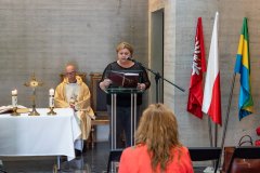 Kobieta czyta słowa Pisma Świętego podczas mszy w krypcie Pomnika Mauzoleum. W tle siedzi ksiądz.  Na stole stoi krzyż pośród dwóch świec.