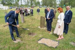 Uczestnicy uroczystości stoją przy alei bohaterów. Mężczyzna sadzi dąb.