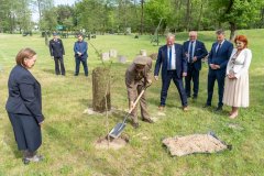 Uczestnicy uroczystości stoją przy alei bohaterów. Mężczyzna w mundurze Lasów Państwowych obsypuje ziemią posadzony przed chwilą dąb.