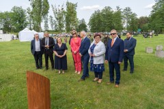 Grupa kilku osób stojących na terenie Fortów Bema przy miejscu posadzenia dębu. Daleko w tle wiele ludzi - uczestników obchodów oraz wysokie drzewa.