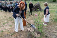 Kobieta wkopuje sadzonkę dębu. W tle kilka innych osób oraz kilkunastu harcerzy i harcerek w dwuszeregu