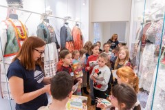 Grupa kilkunastu dzieci ogląda wystawę tradycyjnych kurpiowskich strojów.