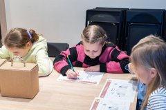 Trzy dziewczynki siedzą przy stole i rozwiązują karty edukacyjne. Na stole stoi tekturowy kuferek. W tle oparte o ścianę plastikowe krzesła